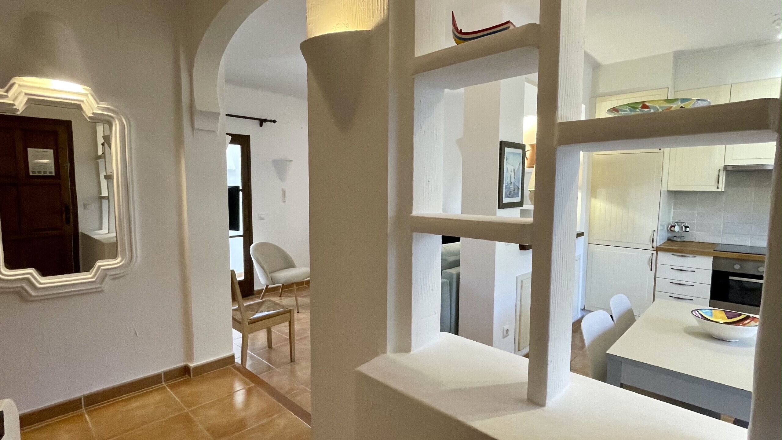 Une cuisine moderne avec des meubles blancs, un coin repas avec une table et des chaises blanches, et un couloir avec une porte blanche et une chaise.