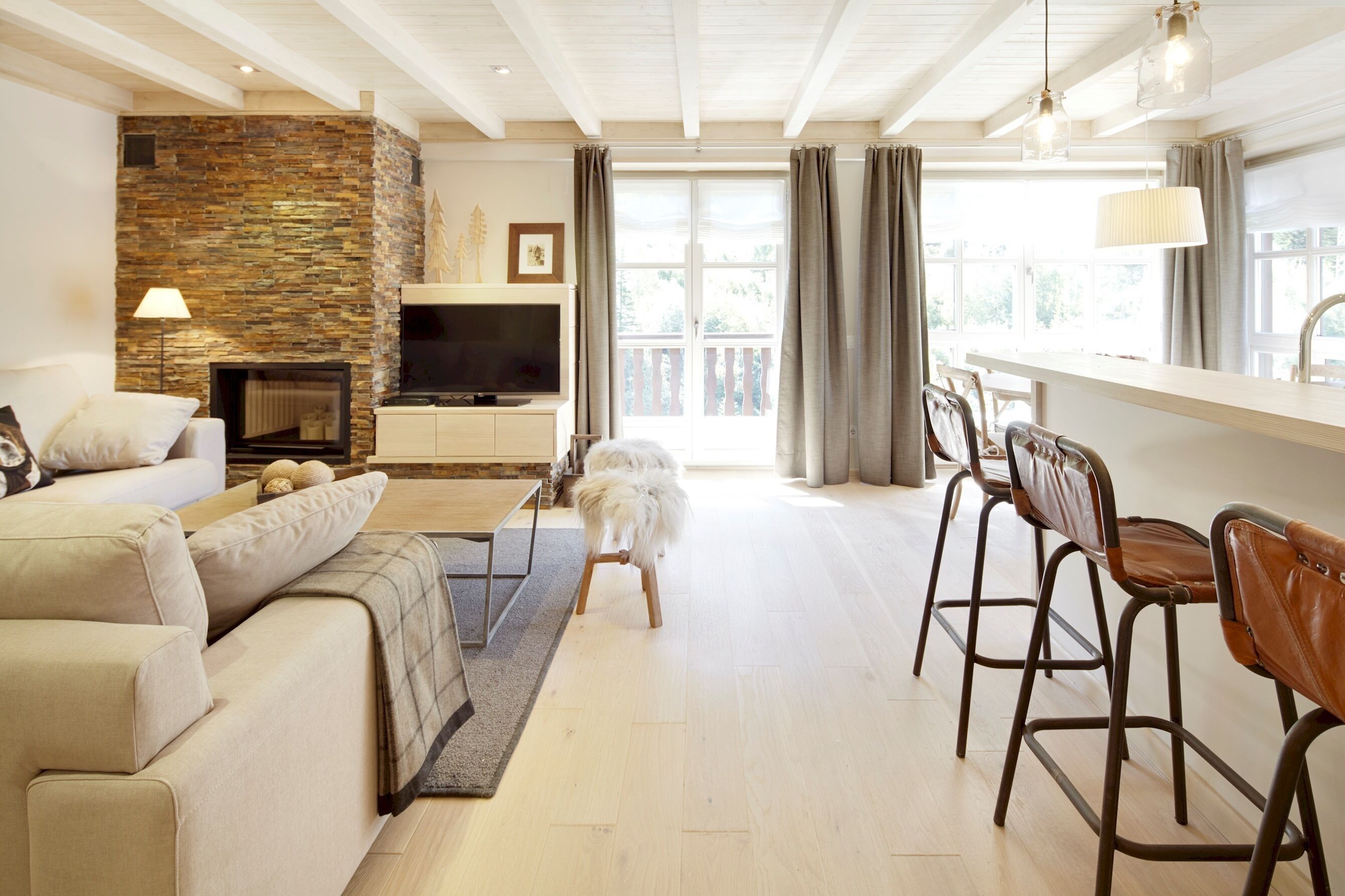 A modern living room with a stone fireplace, a sofa, a dining area with bar stools, and a large window with curtains.