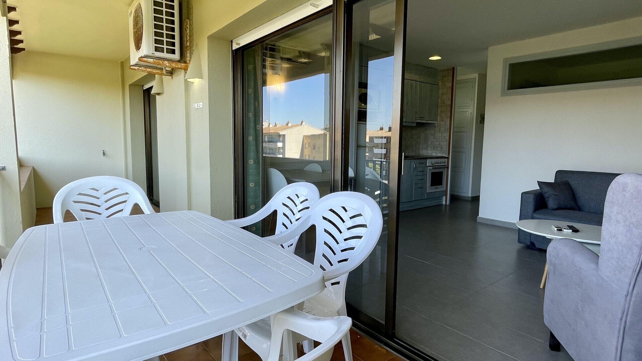 Varanda com mesa e cadeiras, porta de vidro deslizante que dá para uma cozinha e um sofá cinzento na sala de estar.