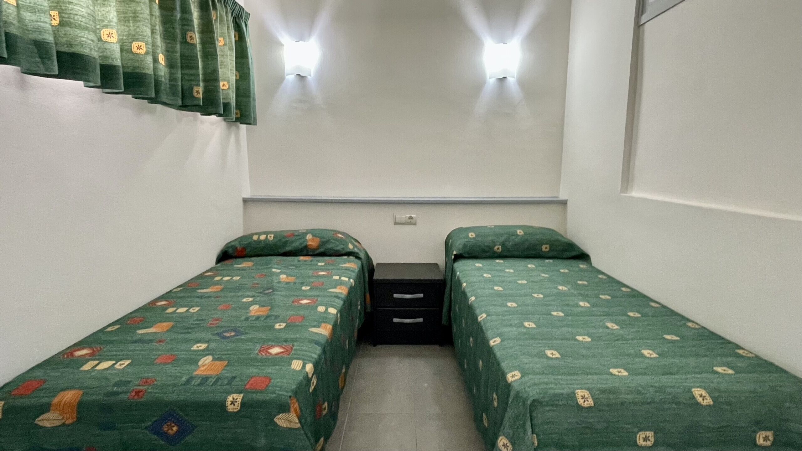 Une petite chambre avec un seul lit, deux lits simples, une petite commode et deux lampes fixées au mur.