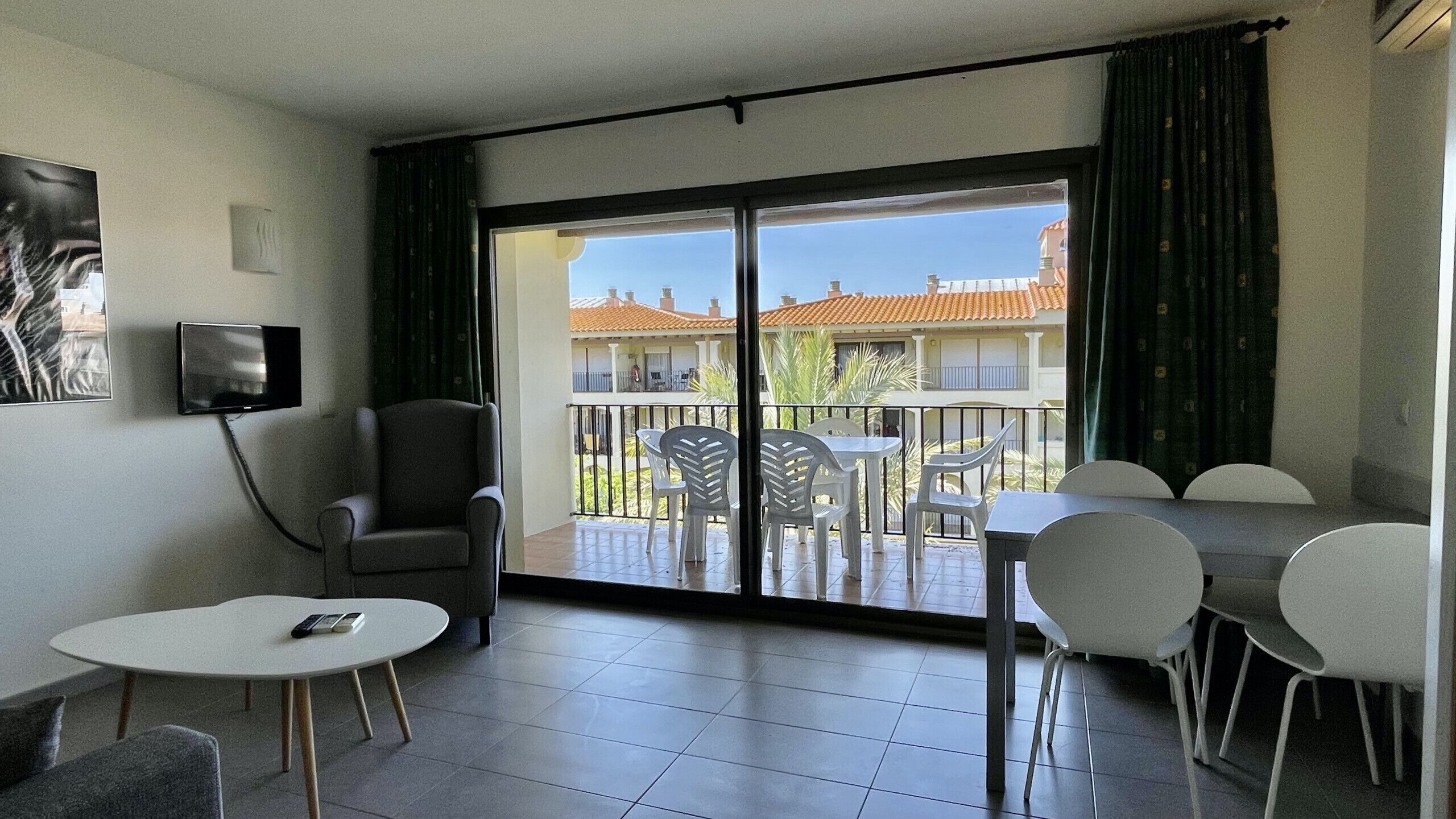 Ein Zimmer mit einer Schiebeglastür, die auf einen Balkon führt, einem Sessel, einem kleinen Tisch und einem an der Wand befestigten Fernseher.