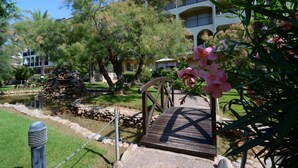 Un puente de madera que conduce a un edificio con una fuente de piedra, rodeado de árboles y un césped bien cuidado.