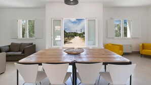 A dining area with a wooden table, white chairs, and a view of a pathway leading to a natural landscape. - Villa Rosalorè by Perle di Puglia (Ugento)