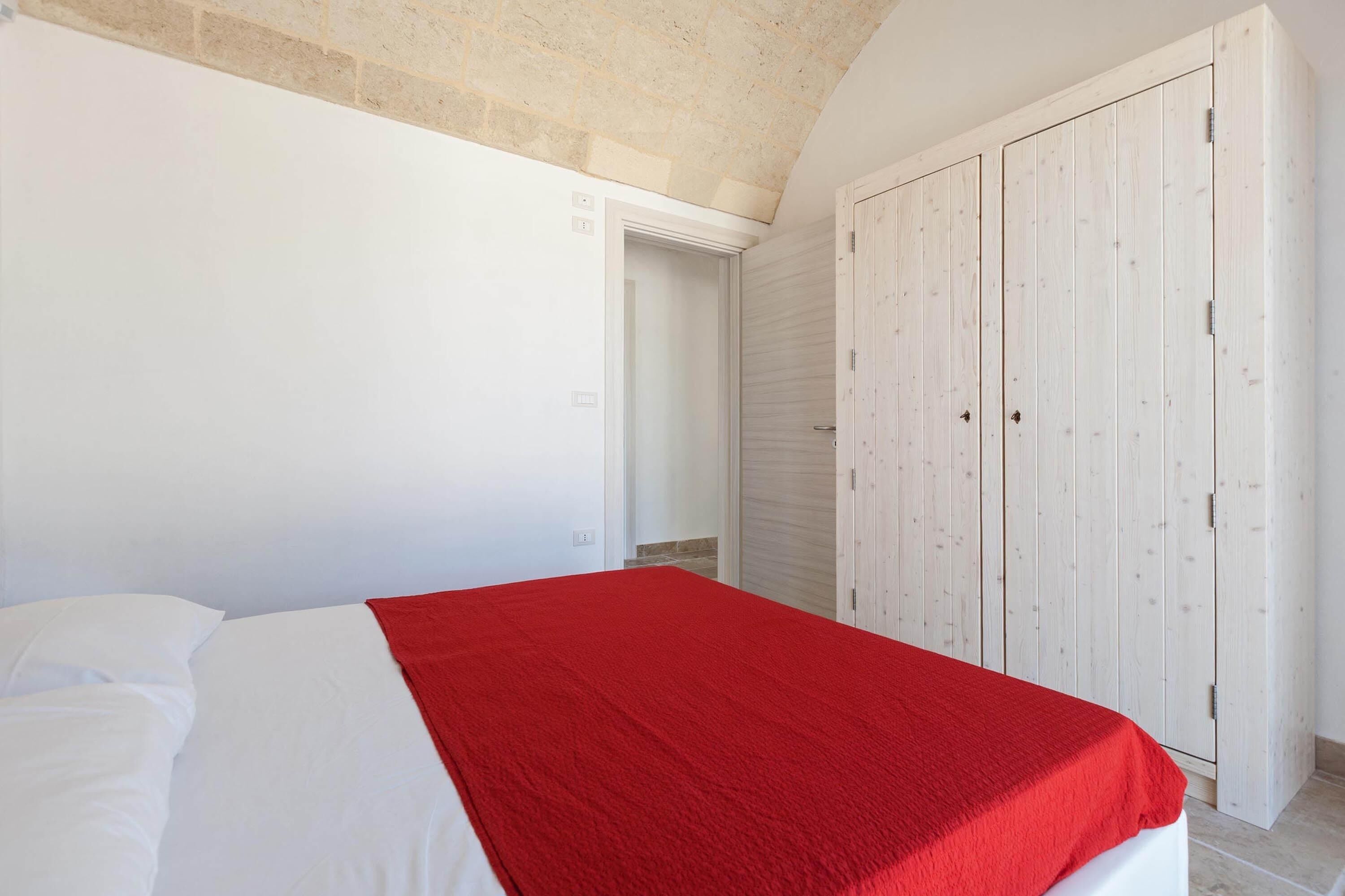 Ein Schlafzimmer mit einem Bett, einer roten Decke, einem weißen Kleiderschrank und einer Tür zu einem anderen Raum.