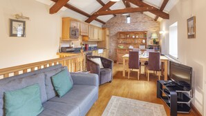 A cozy living room with a sofa, a television, and a kitchen area with wooden cabinets and a stone wall. - The Granary, Luxborough (Luxborough)