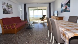 Ein Zimmer mit einem roten Sofa, einem Tisch, einem Fernseher auf einer Holzkommode und Meerblick durch eine Glastür.