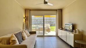 A hotel room with a sofa, a TV, a cabinet, and a sliding glass door leading to an outdoor area with a pool and buildings in the background.