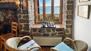 A cozy room with stone walls, a fireplace, wicker chairs, a table with tea set, and a window overlooking the sea. - The Crows Nest, Porlock Weir (Somerset)