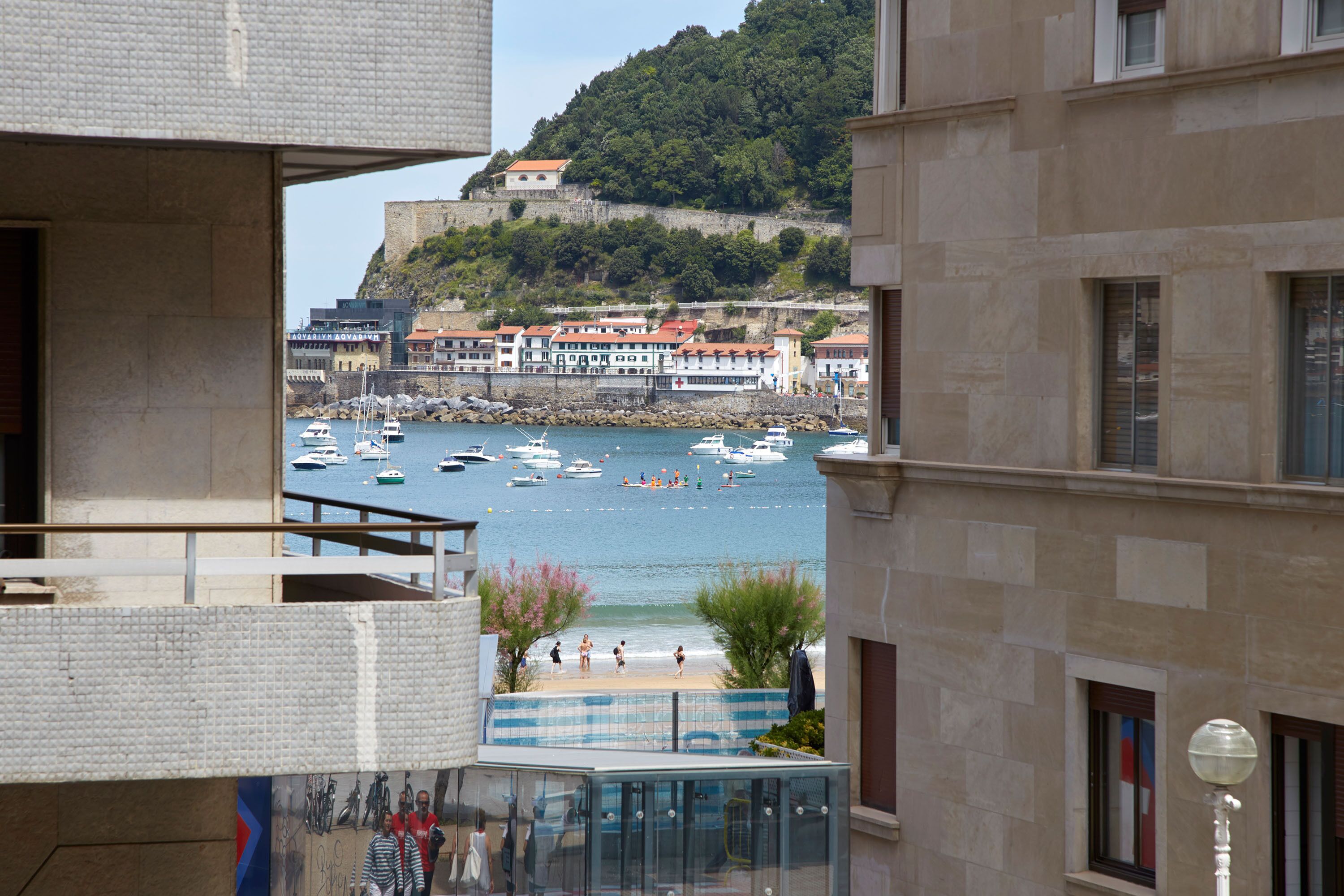 A view of a coastal town with a historic castle on a hill, boats on the water, and buildings with balconies overlooking the sea.