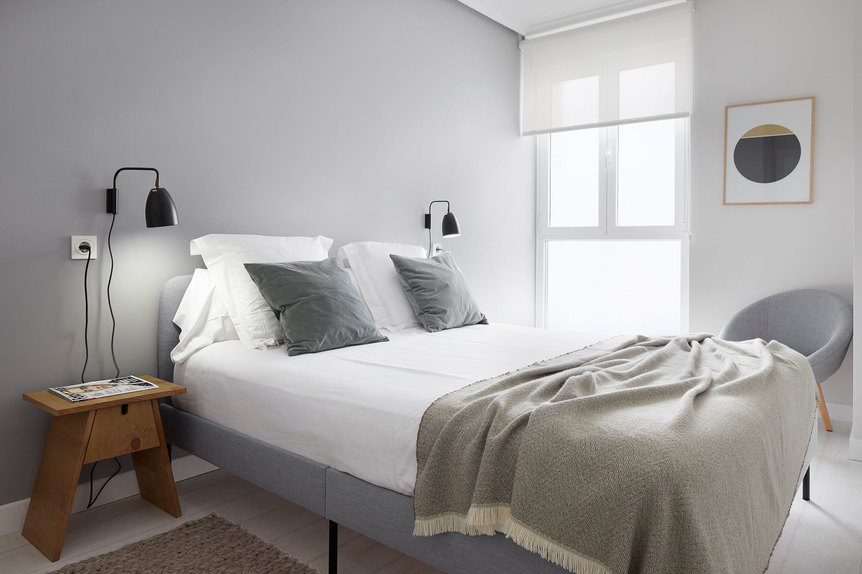A minimalist bedroom with a bed, bedside lamps, a small wooden nightstand, and a chair.
