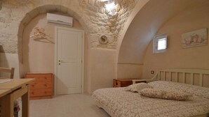 Quarto com arco de pedra, cama com roupa de cama floral, cómoda de madeira e uma pequena mesa.