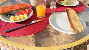 Een ontbijttafel met schalen fruit, toast en sinaasappelsap, op een glazen tafel buiten, met uitzicht op bomen en een gebouw op de achtergrond.