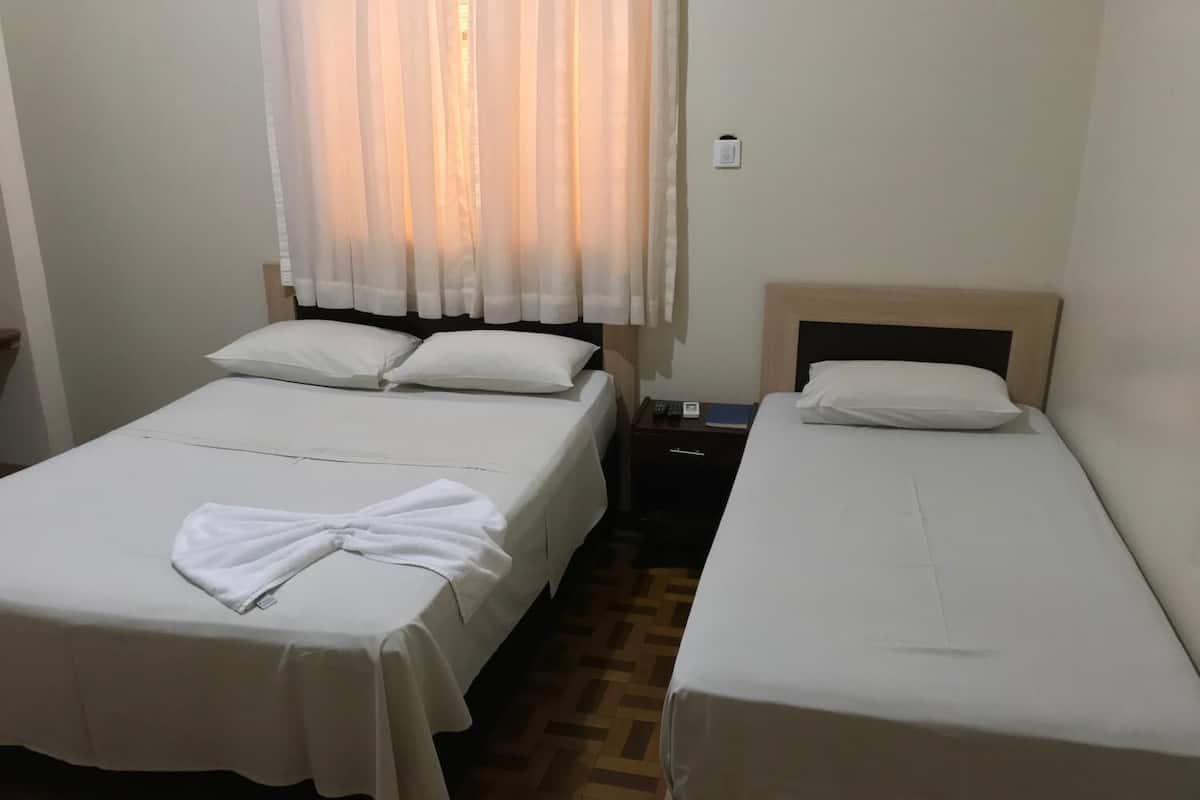 Una habitación con dos camas, cada una con ropa de cama y almohadas blancas, un cabecero de madera, una mesita de noche con un libro y un control remoto, y una ventana con cortinas traslúcidas.