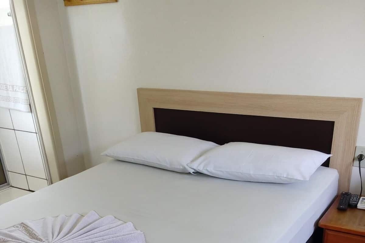 Una cama bien hecha con sábanas blancas y una toalla decorativa, un cabecero de madera, una mesita de noche con un teléfono y una lámpara fijada a la pared.