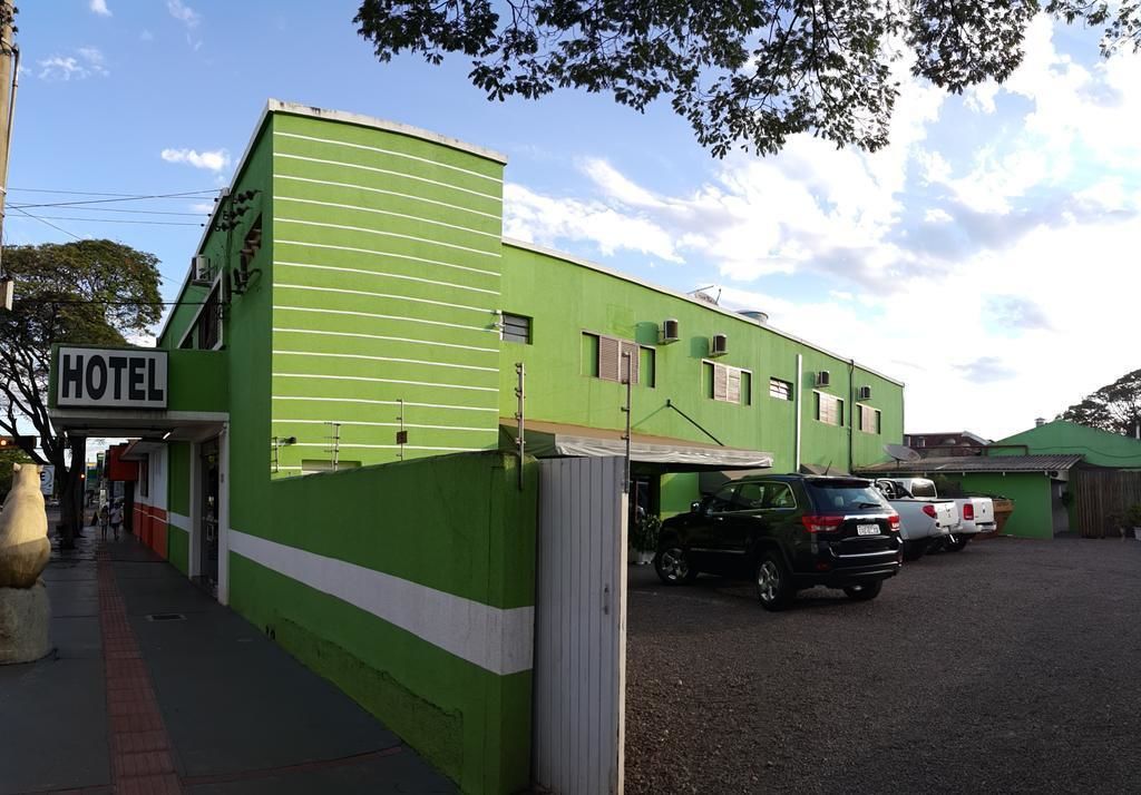Un edificio verde con un cartel que dice 'HOTEL', una zona de estacionamiento con coches y un cielo despejado.