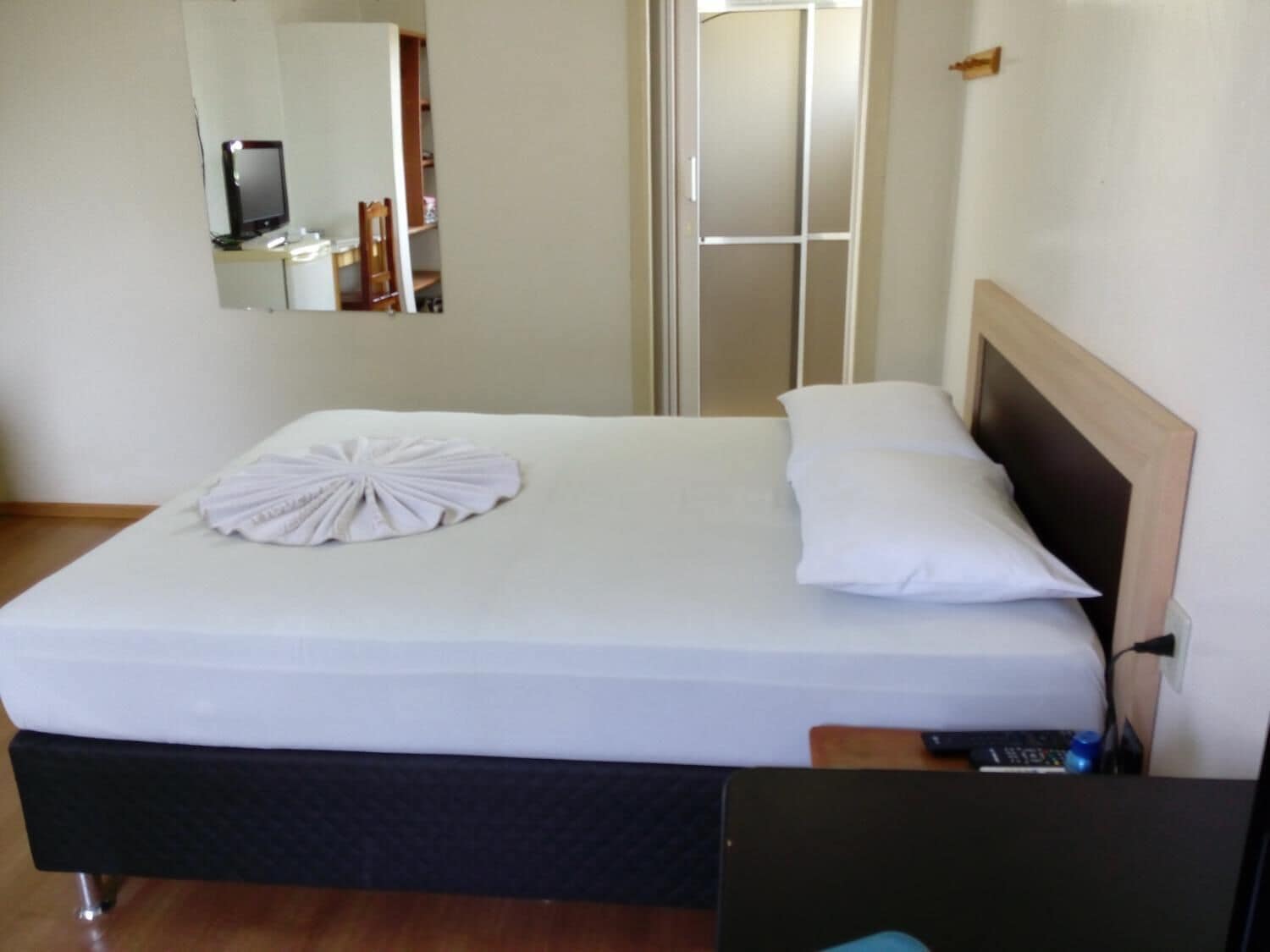 Ein ordentlich bezogenes Bett mit weißer Bettdecke und Kissen, ein Holz-Kopfteil, ein Nachttisch mit Fernbedienung und ein Spiegel an der Wand.