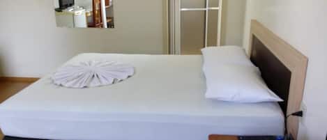 Una cama bien hecha con edredón blanco y almohadas, cabecera de madera, mesita de noche con control remoto y un espejo en la pared.