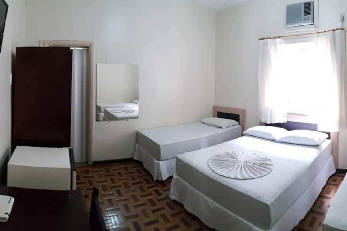 Habitación de hotel con dos camas individuales, una mesita, una silla, un televisor, un espejo, una ventana con cortinas y un aire acondicionado instalado en la pared.