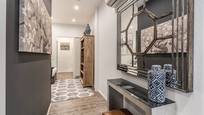 A hallway with a wooden floor, a gray rug, a bench, a console table with decorative items, and a large mirror with a hexagonal frame.