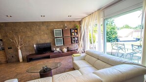 A modern living room with a large beige sofa, a glass coffee table, a flat-screen TV, and a bookshelf.