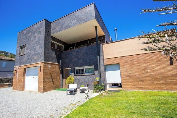 Une maison moderne à deux étages, dotée d’un toit plat, d’un garage et d’un petit patio aménagé avec du mobilier d’extérieur.
