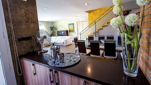 A modern kitchen with a central island, a dining area with a staircase, and a living room with a sofa and a plant.