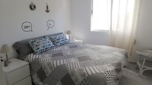 A neatly made bed with patterned bedding, a bedside table with a lamp, and a small round table in a bright room with a window.