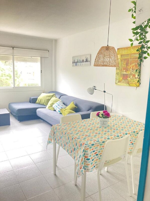 Un salon comprenant un canapé, une table à manger avec des chaises, un luminaire suspendu et une fenêtre avec des stores.