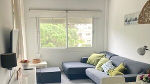 A compact living space with a sofa, a small table, a TV, and a window with a view of greenery.