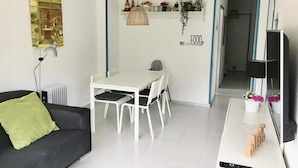 A modern living room with a black sofa, a white dining table with chairs, a hanging light fixture, and a wall-mounted shelf with decorative items.