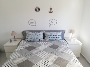 A neatly made bed with patterned pillows, a patchwork quilt, two bedside tables with lamps, and wall decorations including a wreath and an anchor.