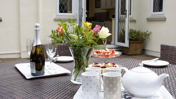 A table set for a meal with a bottle, glasses, a vase with flowers, and a teapot. - Plum Tree Cottage, Porlock (Porlock)