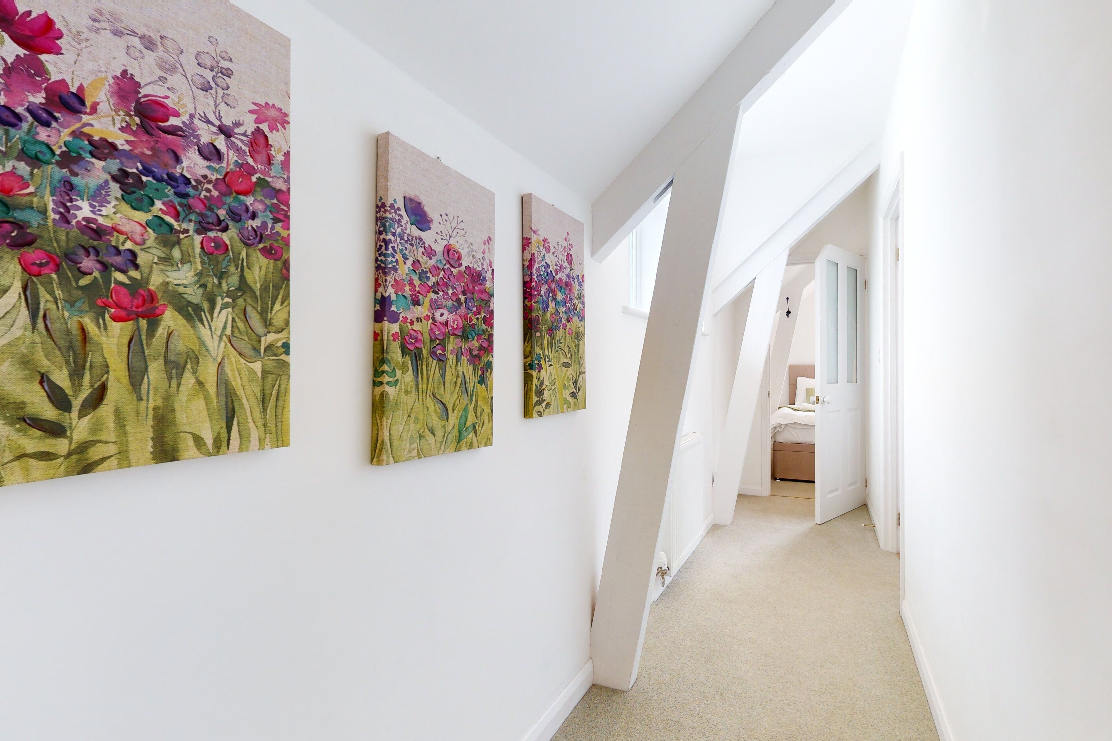 A hallway with three floral paintings on the wall, leading to a bedroom with a bed and a nightstand.