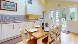 A kitchen with white cabinets, a wooden dining table set for four, and a window with a view of greenery. - Plum Tree Cottage, Porlock (Porlock)