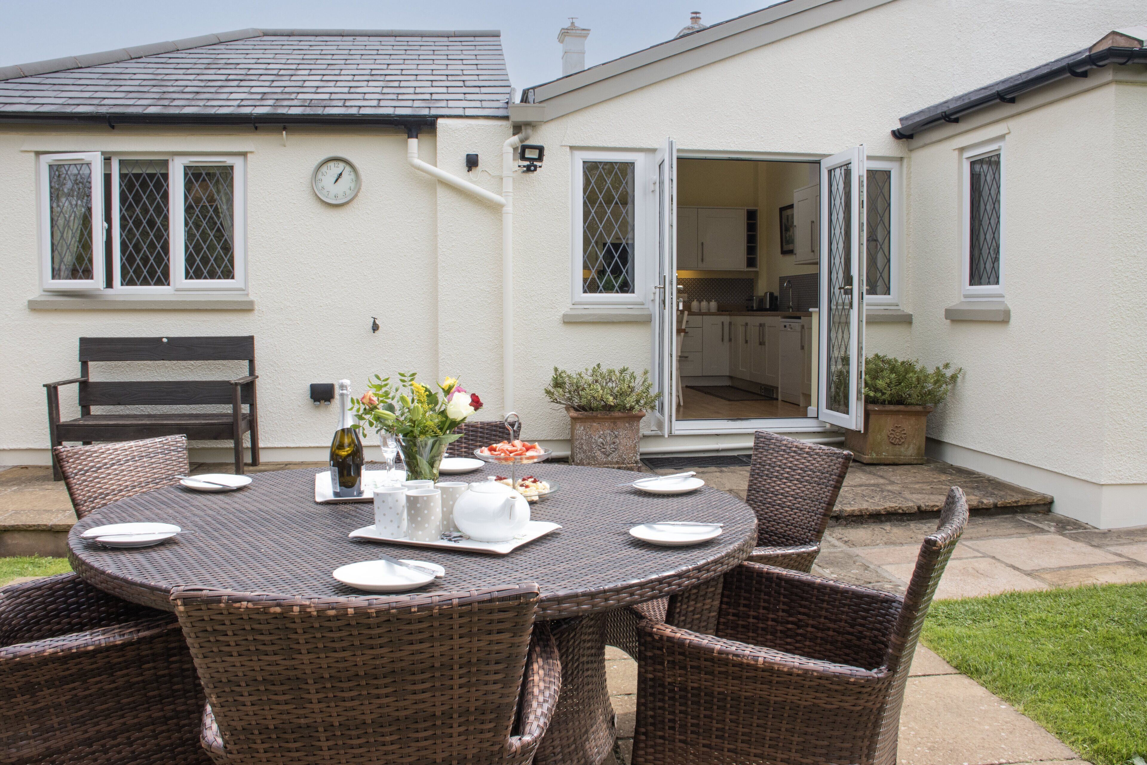 A patio with a round table set for four, surrounded by wicker chairs. A bench is positioned to the left of the table.