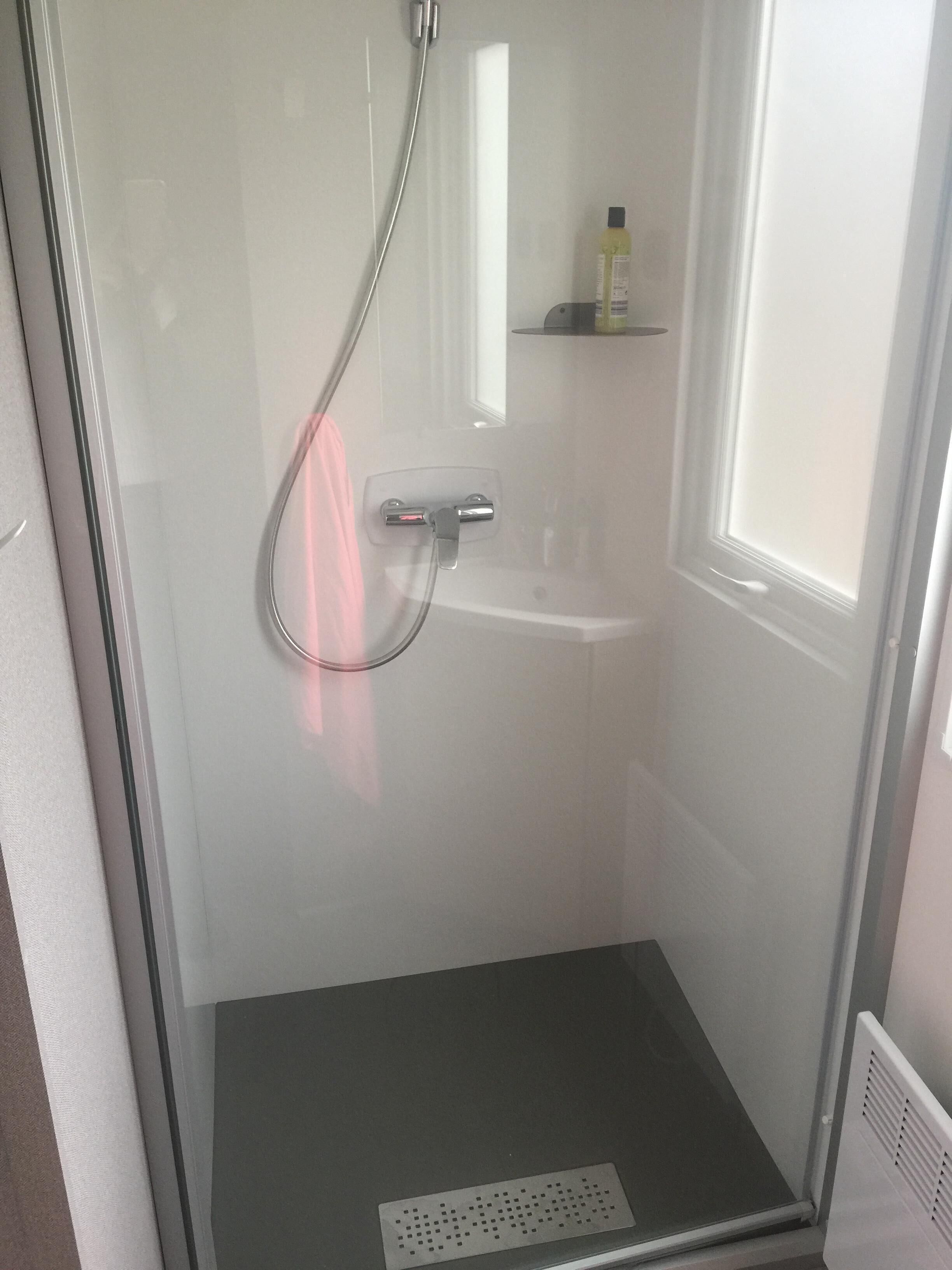 A glass-enclosed shower with a handheld showerhead, a small shelf with toiletries, and a vent on the floor.