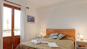 Quarto de hotel com uma cama, uma mesinha de cabeceira, uma luminária, um quadro na parede e uma porta com cortinas.