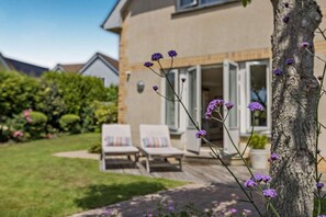 Une maison avec un jardin, deux fauteuils de jardin et des fleurs violettes.