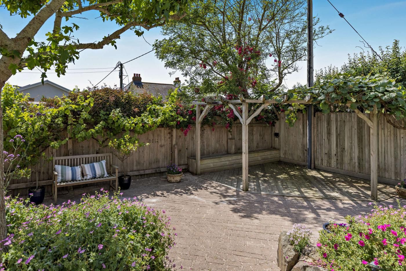 Une pergola en bois ornée de plantes grimpantes, un banc en bois avec des coussins et une terrasse en briques.
