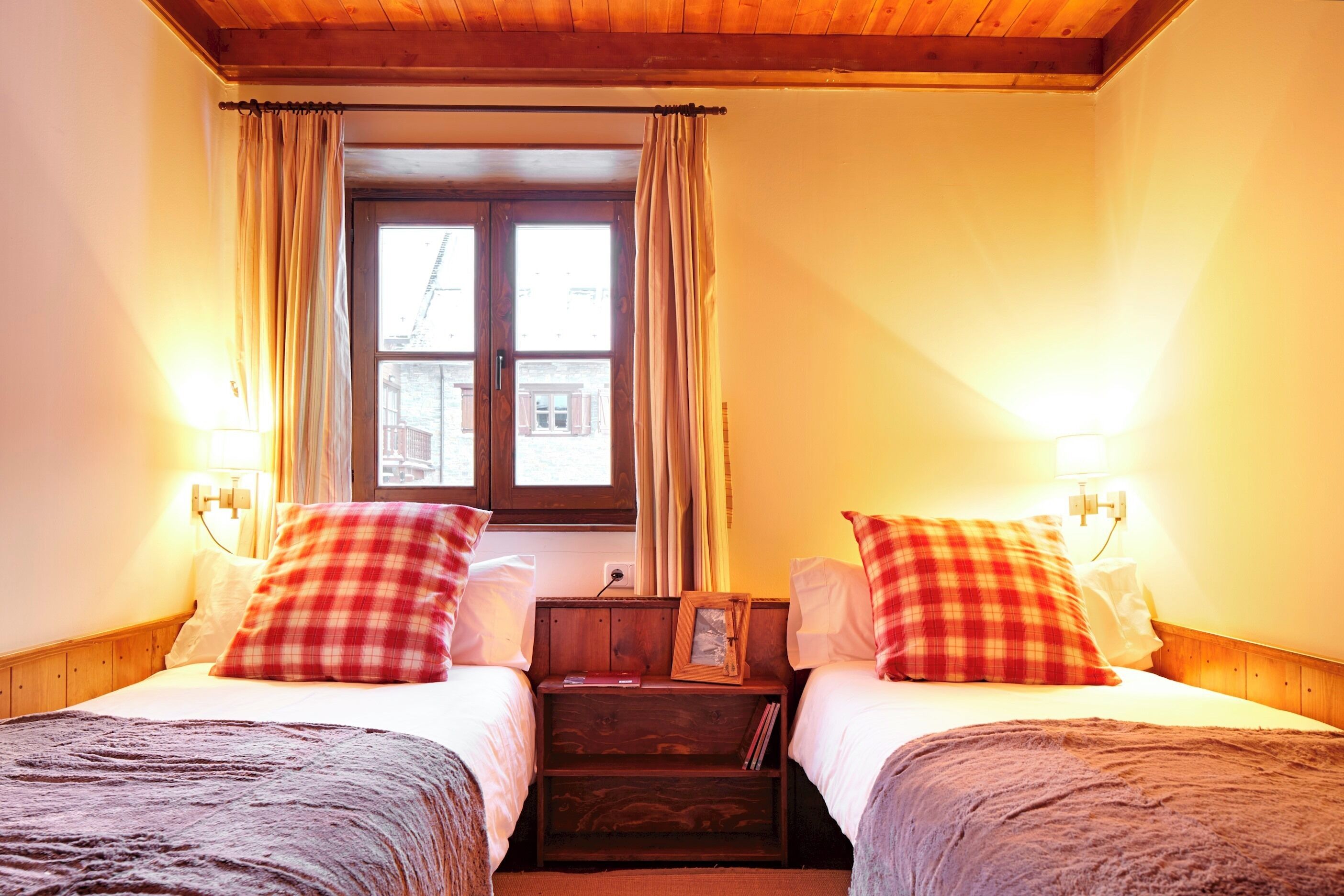 A double room with two beds, each with a red and white checkered pillow, a bedside table with a lamp, and a window with curtains.