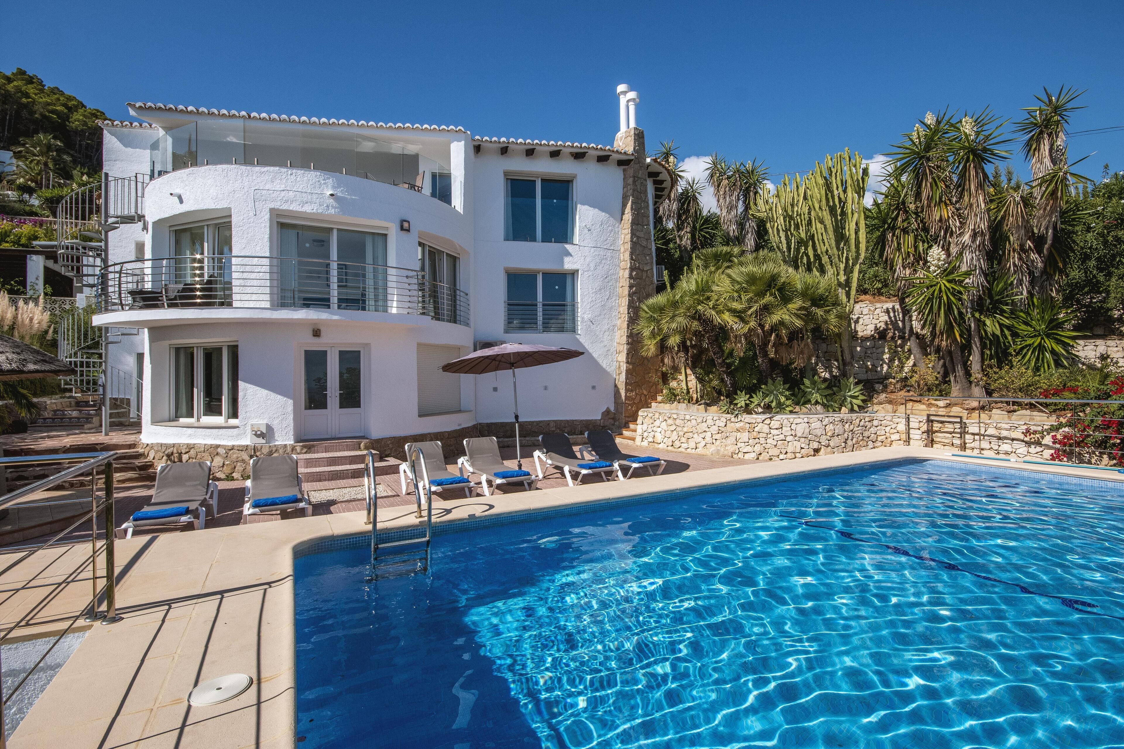 A two-story building with a swimming pool, lounge chairs, and a stone wall.