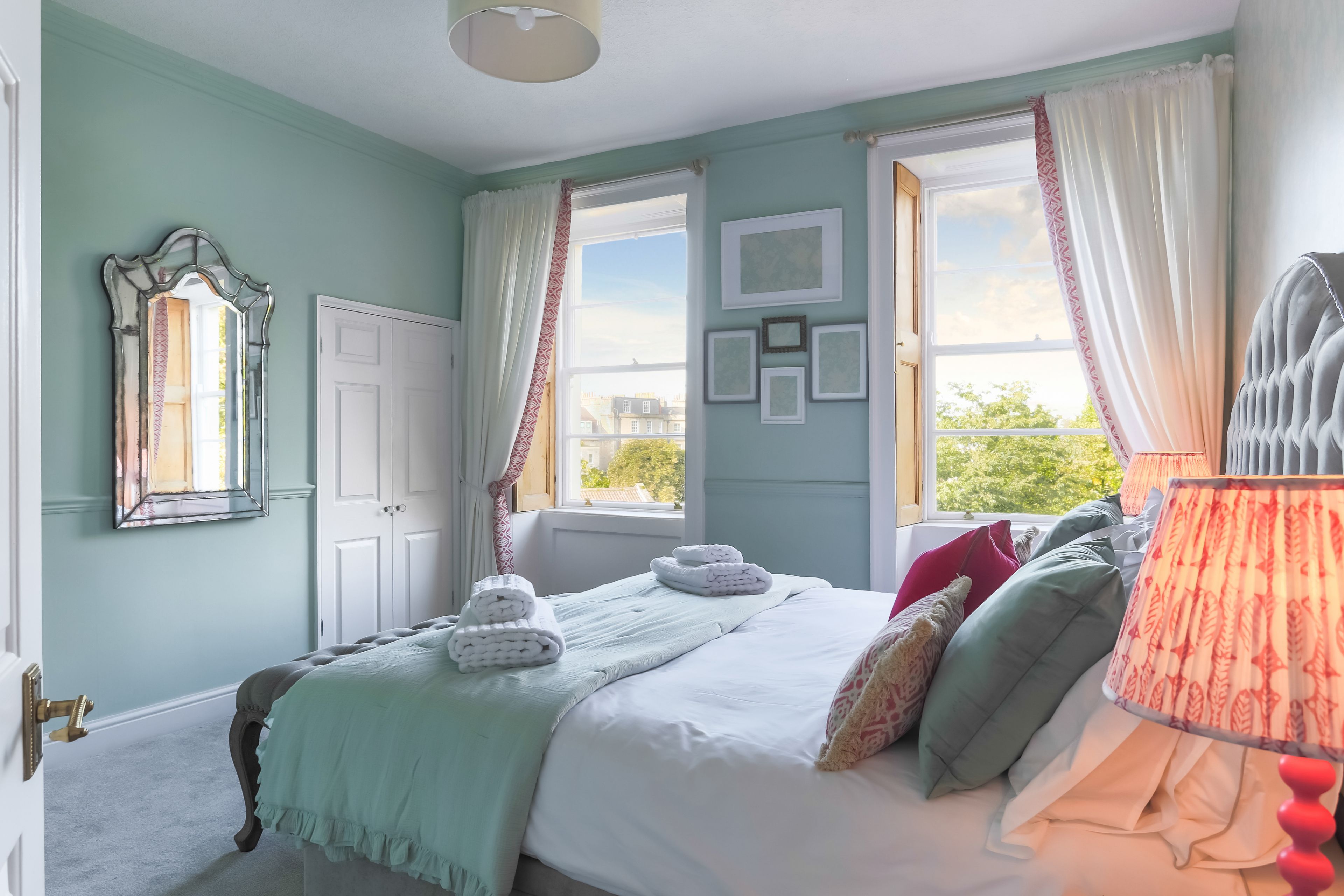 Une chambre à coucher avec un lit, une fenêtre avec des rideaux, un miroir et un mur orné de tableaux.