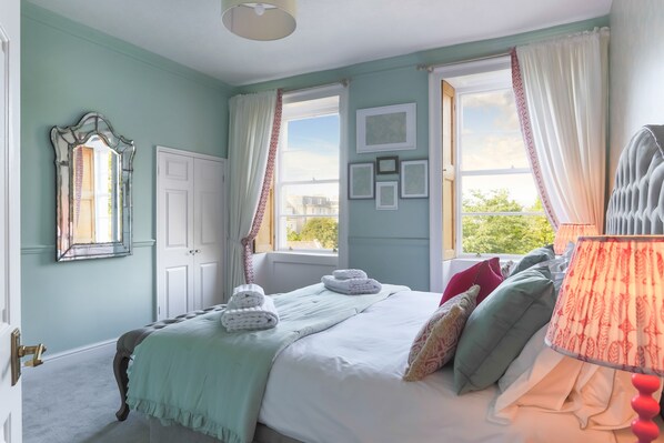 A bedroom with a bed, a window with curtains, a mirror, and a wall with framed pictures.