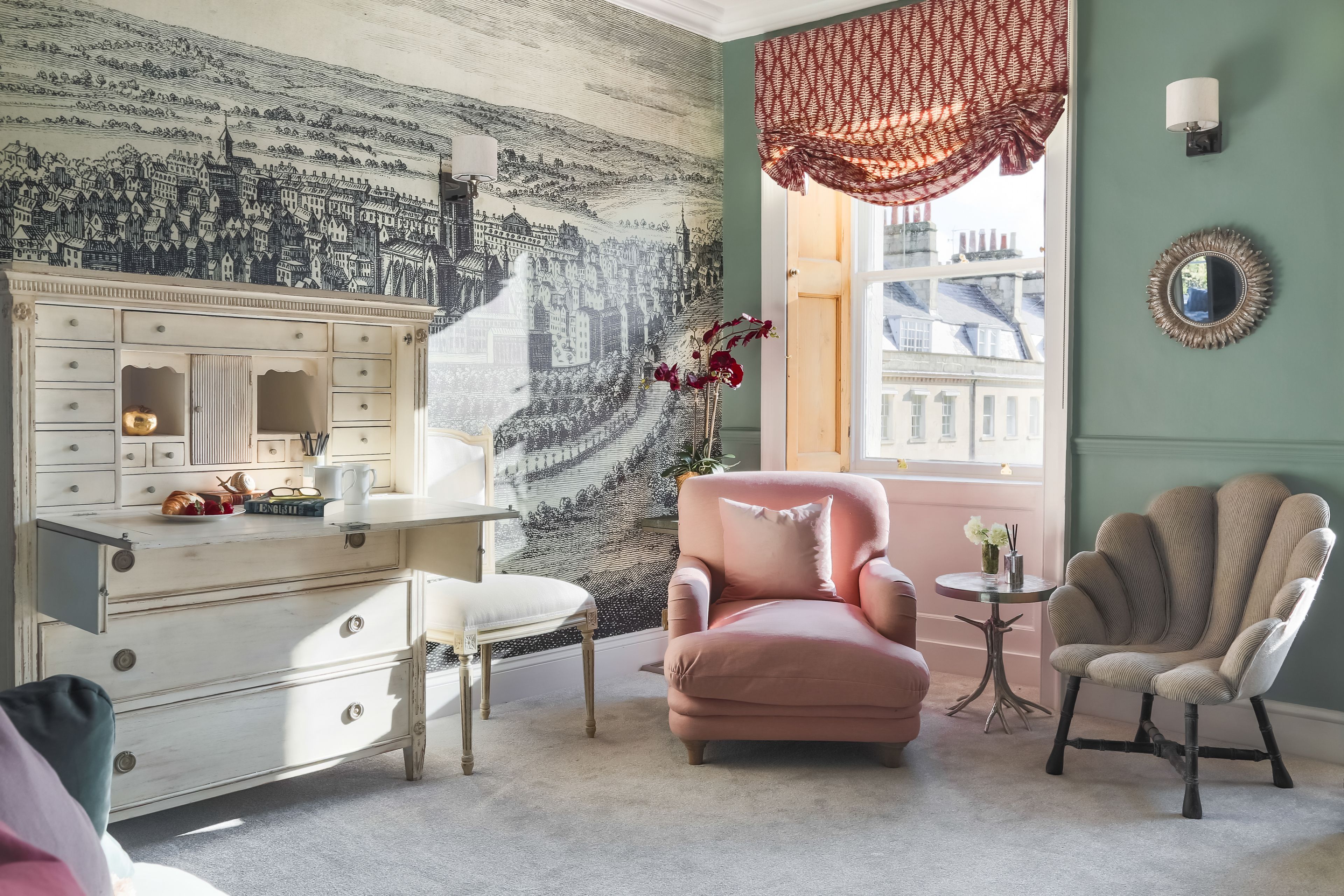 Una habitación con un aparador antiguo, un sillón rosa, una mesa redonda con un jarrón y un ventanal con vistas a edificios.