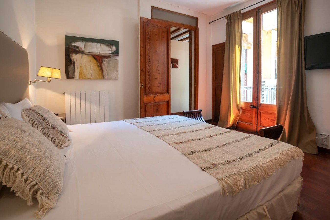 A hotel room with a bed, a bedside lamp, a painting on the wall, a wooden door, and a window with curtains.
