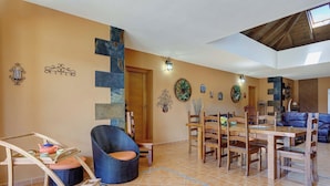 A spacious living area with a dining table, chairs, a blue wicker chair, a wooden cart, and a tiled floor.