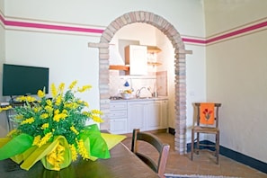 A dining area with a brick archway leading to a kitchen, a table with a bouquet of yellow flowers, and a chair with a painting.