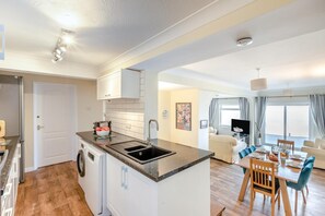 A modern kitchen with a central island, a dining area with a table and chairs, and a living room with a sofa and TV. - 3 Bed in Clifton Court (oc-fl8) (Croyde)