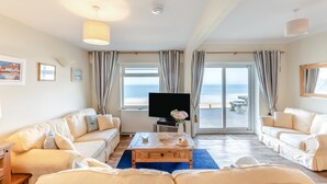 A hotel room with a large window, a flat-screen TV, a wooden coffee table, and two sofas. - 3 Bed in Clifton Court (oc-fl8) (Croyde)