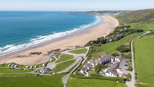 An aerial view of a coastal area with a sandy beach, a small settlement with houses and parked cars, and green fields. - 3 Bed in Clifton Court (oc-fl8) (Croyde)
